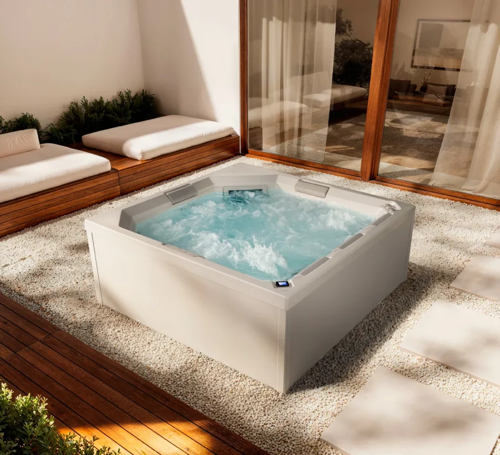 Vierkante witte spa met bruisend water, vrijstaand op een grindbed naast een houten vlonder en een glazen pui, de Aquadvance 210 van Gruppo Treesse